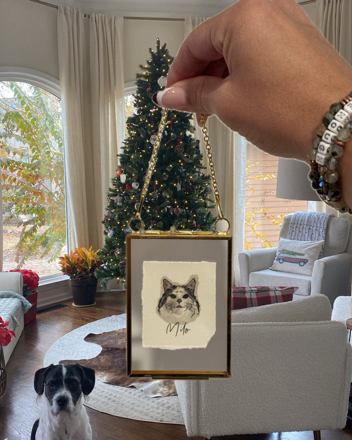 A hand holds up a small framed custom pet portrait featuring a black and white sketch of a dog on textured cream paper, enclosed in a gold frame with a gold hanging loop. A black and white dog sits on the wooden floor in the background of a cozy living room with a Christmas tree.