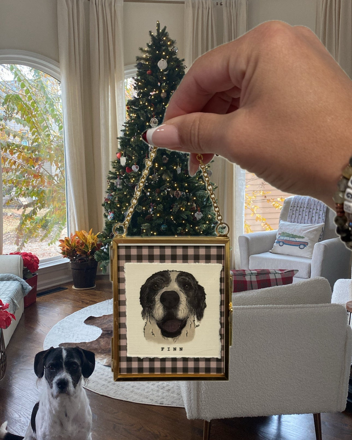 A hand holds a small framed Christmas ornament featuring a black and white dog portrait with the name 'PIMU' below, while a real dog sits on the floor in a decorated living room with a Christmas tree in the background.