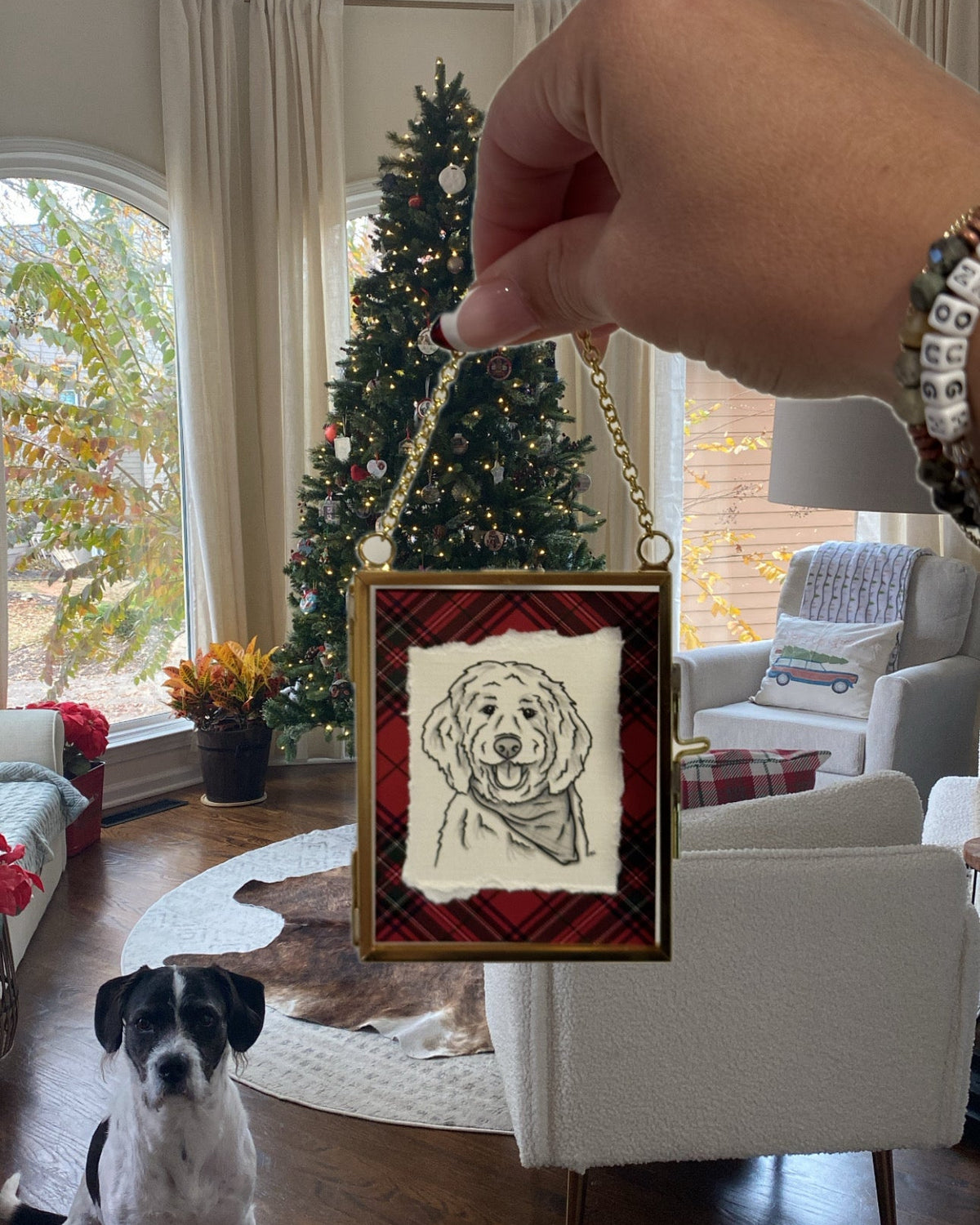 A hand holds up a square Christmas ornament featuring a black-and-white sketch of a dog on a cream background, surrounded by a red plaid border and gold frame. A black-and-white dog sits on a hardwood floor in a decorated living room with a Christmas tree in the background.