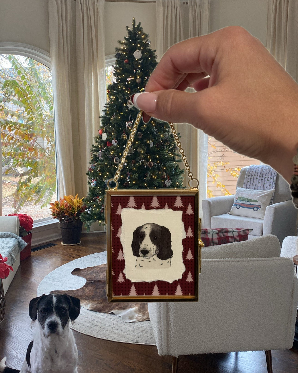 A hand holds a small square Christmas ornament featuring a black and white dog portrait with a red patterned border. A real dog sits on the hardwood floor in a decorated living room with a Christmas tree, chairs, and windows in the background.